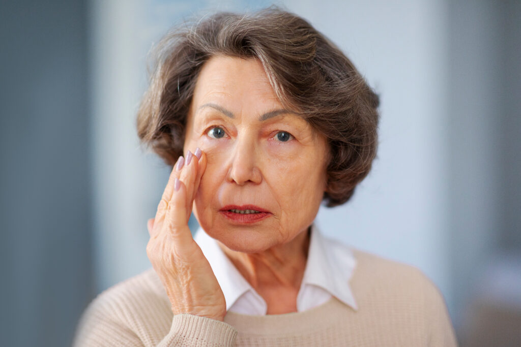 A senior woman touching her cheek and caring for her eye, highlighting the importance of cataract symptoms awareness and eye health.