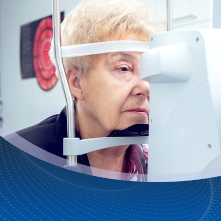 Elderly woman undergoing an eye exam with an ophthalmologist for cataract treatment. She rests her chin on a machine to keep her head steady during the examination.