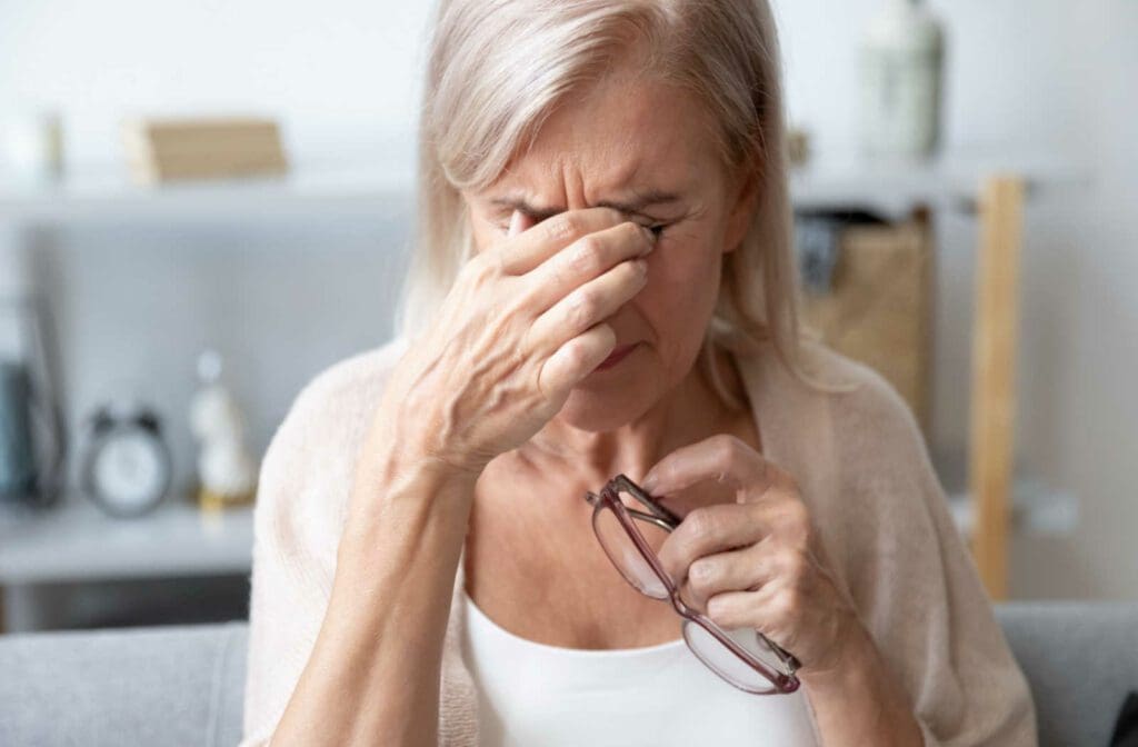 An older woman rubs her eyes as she is experiencing a cloudy vision post her cataract surgery while holding a pair of glasses in her other hand.
