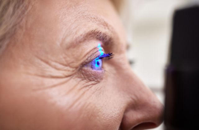 Image of a senior woman having her eyes examined by an ophthalmologist. The woman is sitting in an exam chair and looking towards the camera. The image suggests that the woman is possibly covered by a Medicare plan for her cataract surgery.