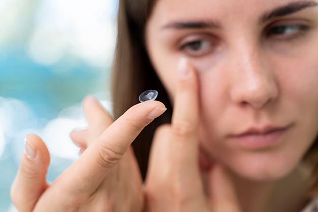 Close-up of a young woman's hand holding a contact lens on her index finger. She appears to be concentrating on placing the lens in her eye.